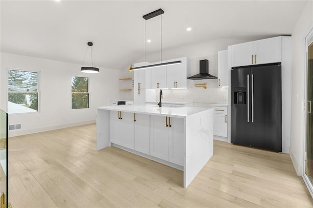 kitchen with wall chimney exhaust hood, black refrigerator with ice dispenser, light hardwood / wood-style floors, a kitchen island with sink, and white cabinets