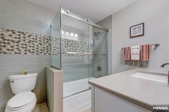 full bathroom featuring toilet, combined bath / shower with glass door, tile walls, wood-type flooring, and vanity