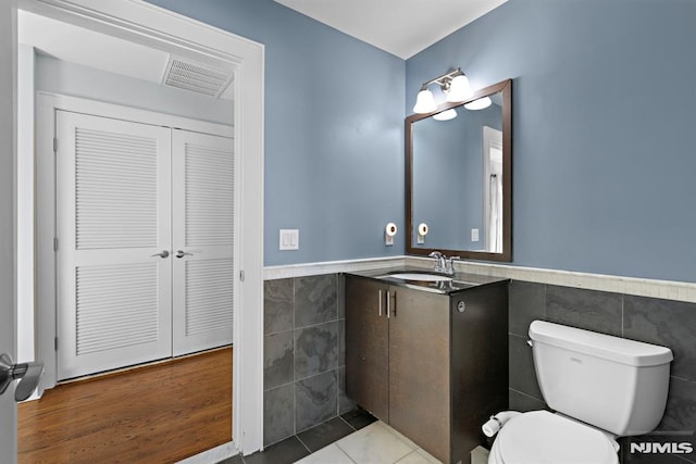 half bathroom with toilet, vanity, visible vents, tile walls, and wainscoting