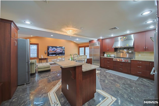 kitchen featuring appliances with stainless steel finishes, wall chimney exhaust hood, a kitchen island with sink, sink, and a breakfast bar area