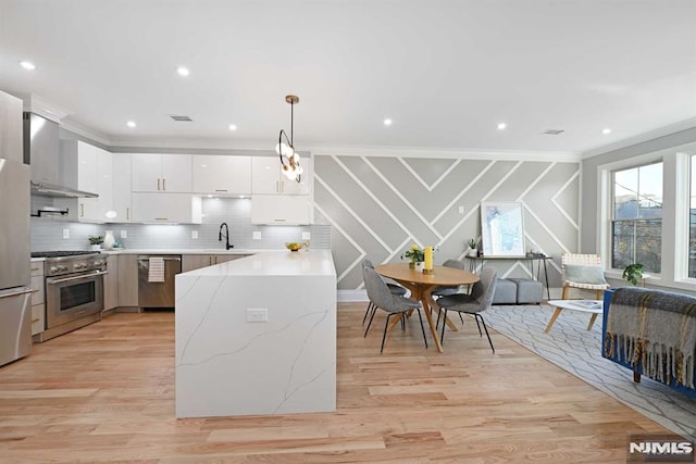 kitchen featuring wall chimney exhaust hood, stainless steel appliances, crown molding, white cabinets, and hanging light fixtures