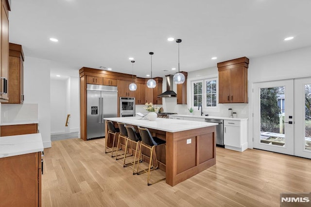 kitchen with wall chimney exhaust hood, appliances with stainless steel finishes, pendant lighting, a kitchen island, and light wood-type flooring