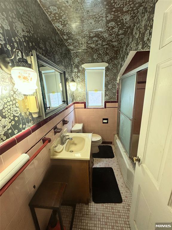 full bathroom with tile patterned flooring, enclosed tub / shower combo, toilet, vanity, and tile walls