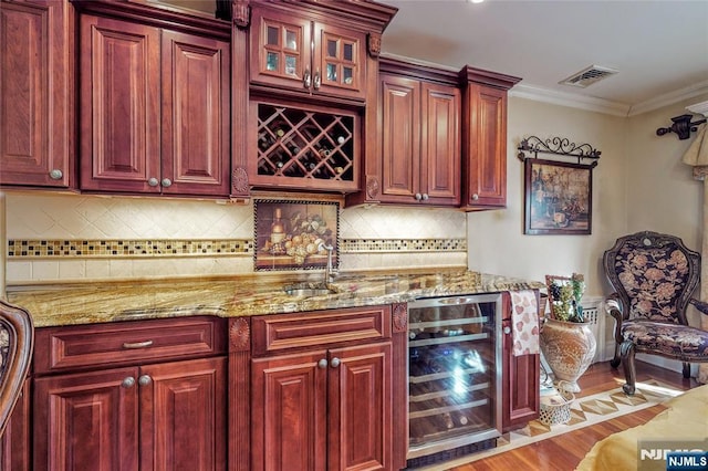 bar with backsplash, wine cooler, ornamental molding, light stone counters, and light wood-type flooring