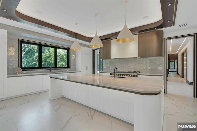 kitchen featuring decorative light fixtures, a raised ceiling, white cabinetry, and an island with sink