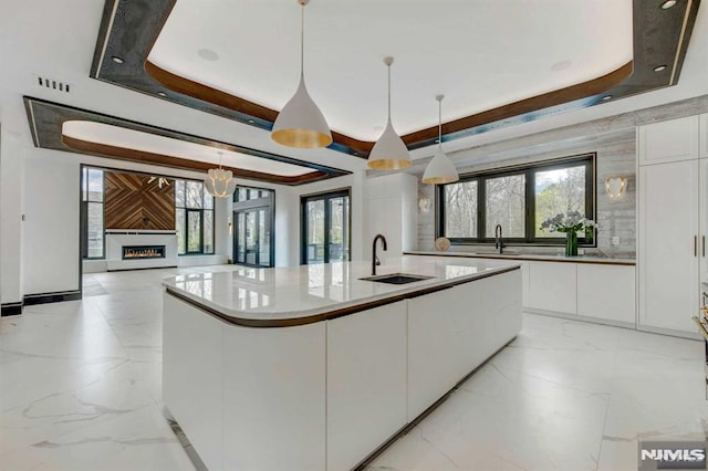 kitchen featuring a tray ceiling, sink, decorative light fixtures, a center island with sink, and white cabinetry