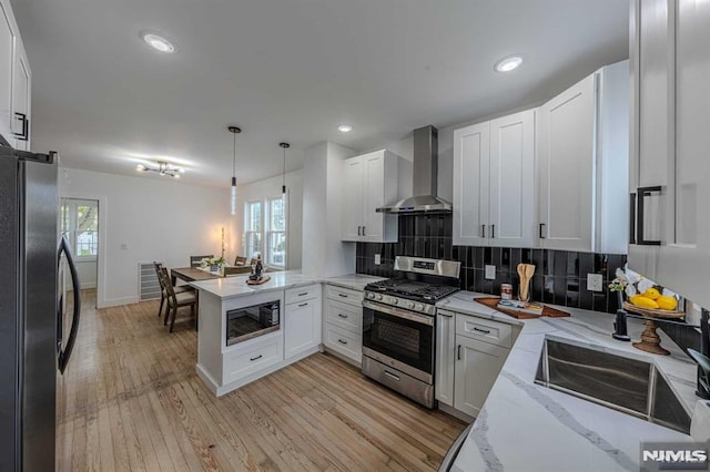 kitchen with kitchen peninsula, appliances with stainless steel finishes, wall chimney range hood, decorative light fixtures, and white cabinets