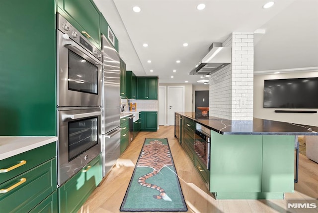 kitchen featuring wall chimney range hood, green cabinetry, decorative backsplash, double oven, and a kitchen bar