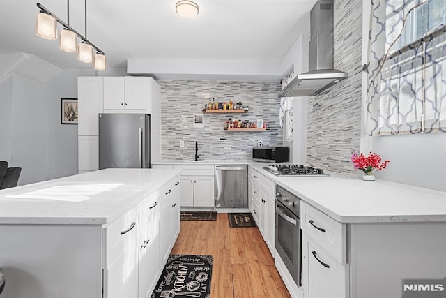 kitchen with white cabinets, wall chimney range hood, hanging light fixtures, appliances with stainless steel finishes, and tasteful backsplash