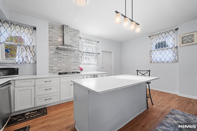 kitchen with appliances with stainless steel finishes, wall chimney range hood, decorative light fixtures, white cabinetry, and a kitchen island