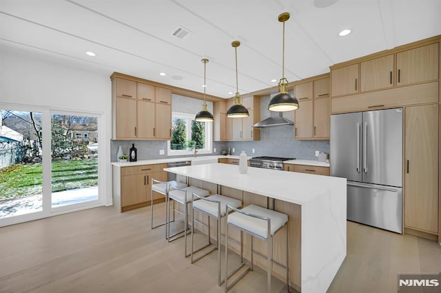 kitchen featuring light brown cabinetry, wall chimney exhaust hood, stainless steel appliances, pendant lighting, and a center island