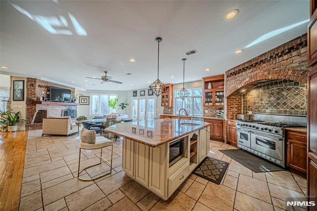 kitchen featuring built in microwave, light stone counters, decorative light fixtures, range with two ovens, and a kitchen island with sink