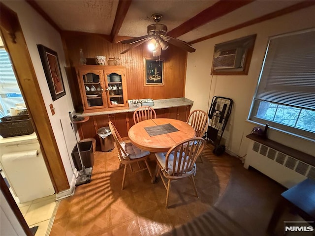 dining room with wooden walls, a wall unit AC, ceiling fan, radiator, and beam ceiling