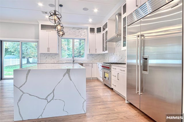 kitchen with light stone countertops, high quality appliances, decorative light fixtures, wall chimney range hood, and white cabinets