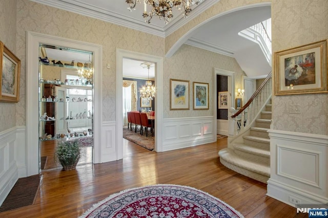entrance foyer featuring wood finished floors, wallpapered walls, arched walkways, a chandelier, and ornamental molding
