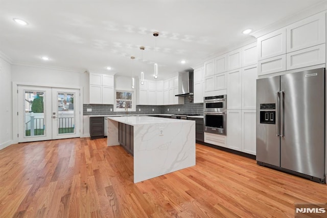 kitchen with appliances with stainless steel finishes, a kitchen island, white cabinetry, wall chimney range hood, and decorative light fixtures