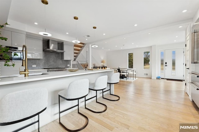 kitchen with backsplash, wall chimney range hood, pendant lighting, light wood-type flooring, and a kitchen breakfast bar