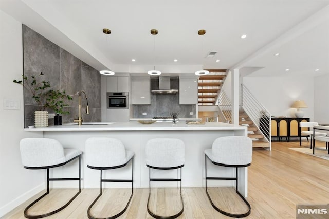 kitchen with decorative light fixtures, wall chimney range hood, sink, stainless steel oven, and light wood-type flooring