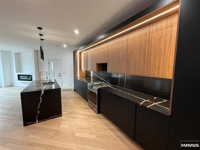 kitchen with sink, hanging light fixtures, stainless steel gas range oven, light hardwood / wood-style flooring, and an island with sink