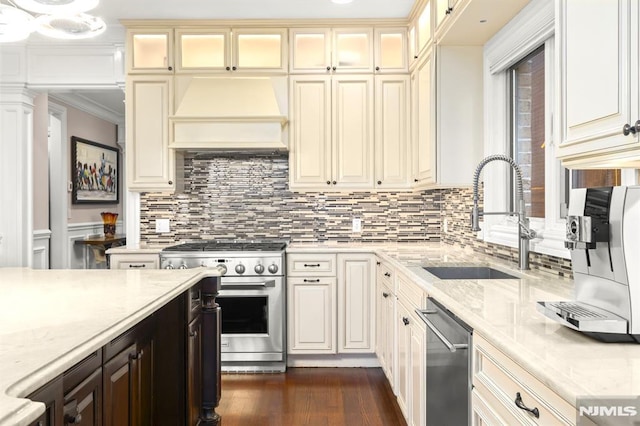 kitchen featuring light stone counters, premium range hood, cream cabinets, stainless steel appliances, and sink