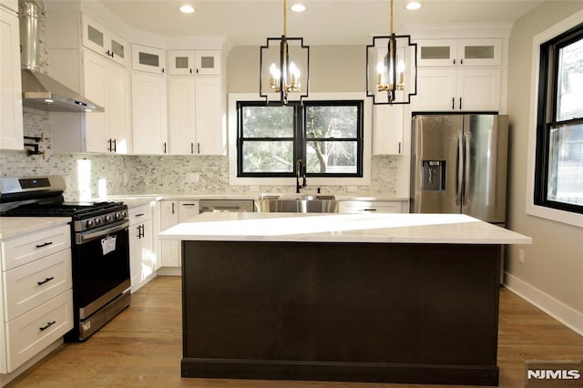 kitchen with wall chimney exhaust hood, stainless steel appliances, a kitchen island, decorative light fixtures, and white cabinets