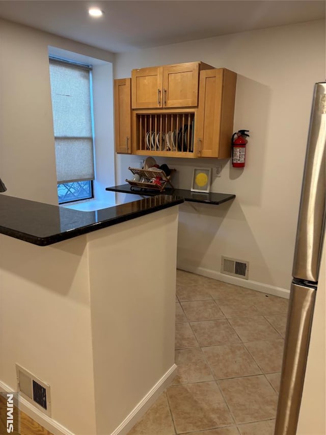 kitchen with stainless steel refrigerator, kitchen peninsula, dark stone countertops, and light tile patterned floors