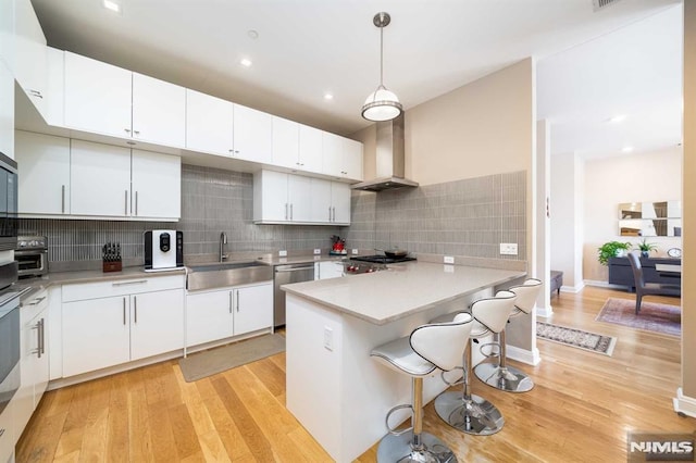 kitchen featuring a kitchen breakfast bar, hanging light fixtures, white cabinets, wall chimney range hood, and sink