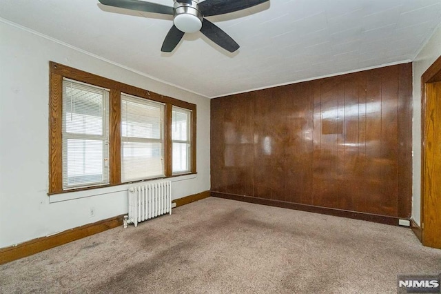 carpeted empty room with ceiling fan, wooden walls, crown molding, and radiator heating unit