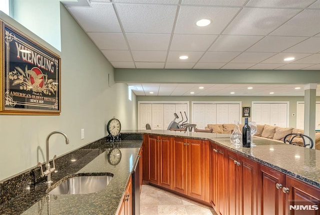 kitchen with sink, light tile patterned flooring, kitchen peninsula, a paneled ceiling, and dark stone counters