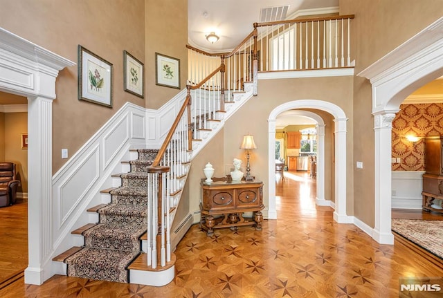 staircase with a towering ceiling, decorative columns, ornamental molding, and parquet flooring