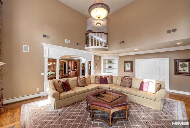 living room with baseboard heating, hardwood / wood-style floors, a towering ceiling, built in features, and decorative columns