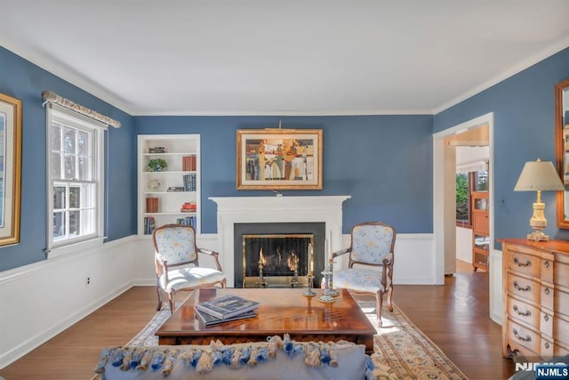 sitting room with built in shelves, crown molding, a wainscoted wall, a lit fireplace, and wood finished floors