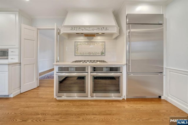 kitchen with ornamental molding, custom range hood, white cabinets, built in appliances, and light wood-type flooring