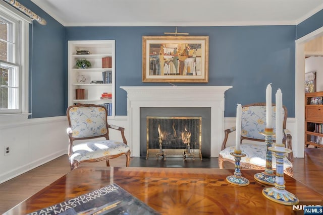 sitting room with built in features, wood finished floors, a wainscoted wall, ornamental molding, and a lit fireplace