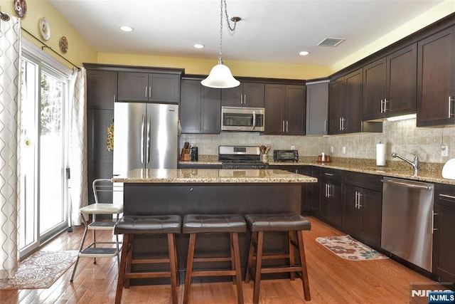 kitchen featuring light stone counters, decorative light fixtures, dark brown cabinets, appliances with stainless steel finishes, and a kitchen island