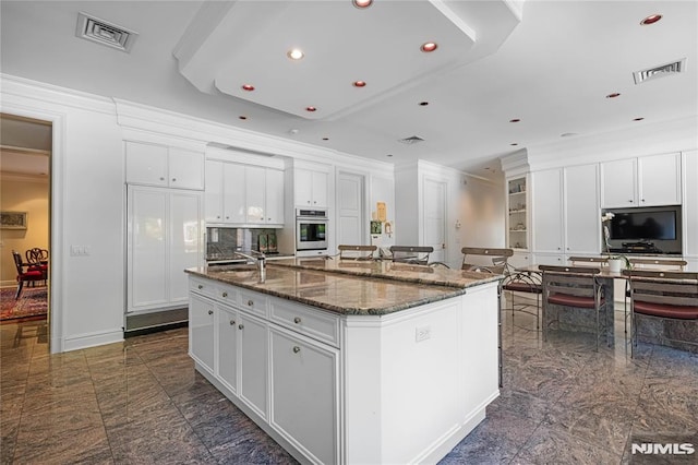 kitchen featuring a kitchen island with sink, dark stone counters, crown molding, and white cabinetry