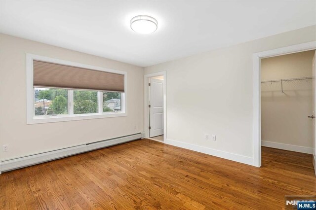 unfurnished bedroom with wood finished floors, baseboards, a closet, a baseboard heating unit, and a spacious closet