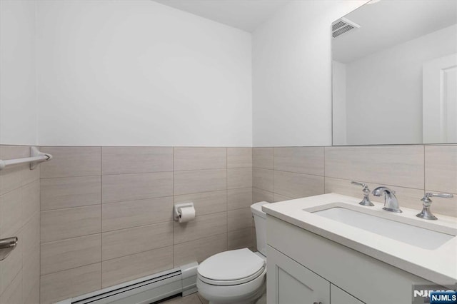 half bath with visible vents, vanity, toilet, tile walls, and a baseboard heating unit