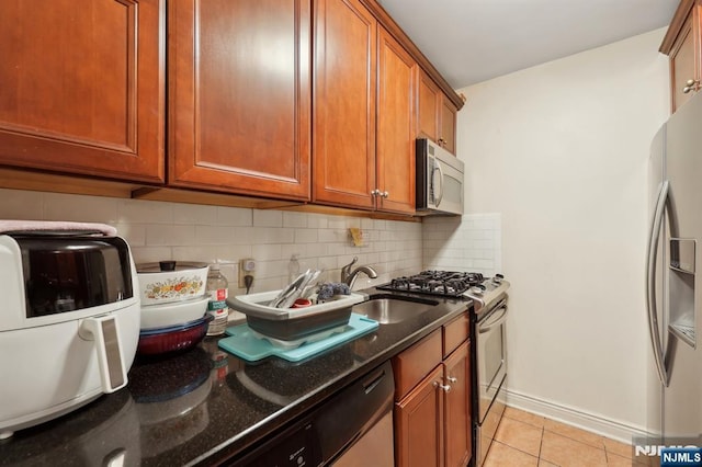 kitchen featuring light tile patterned floors, appliances with stainless steel finishes, decorative backsplash, brown cabinetry, and dark stone countertops