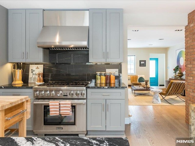 kitchen featuring stainless steel range, decorative backsplash, dark stone counters, wall chimney exhaust hood, and light hardwood / wood-style flooring