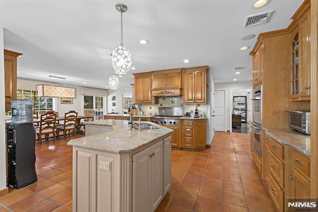 kitchen featuring stainless steel double oven, sink, hanging light fixtures, light stone counters, and a center island with sink