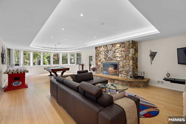 living room featuring a raised ceiling, a stone fireplace, pool table, and light hardwood / wood-style flooring