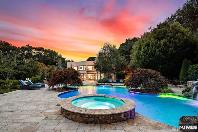 pool at dusk featuring pool water feature, a patio, and an in ground hot tub