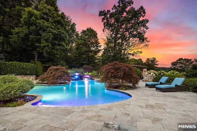 pool at dusk featuring pool water feature and a patio