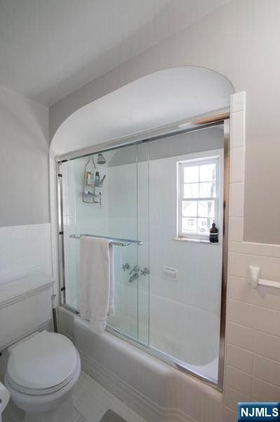 bathroom with toilet, bath / shower combo with glass door, and tile walls