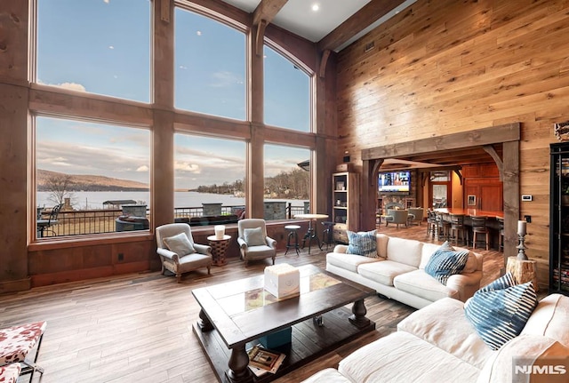 living room with a towering ceiling, wooden walls, light hardwood / wood-style floors, a water view, and beam ceiling