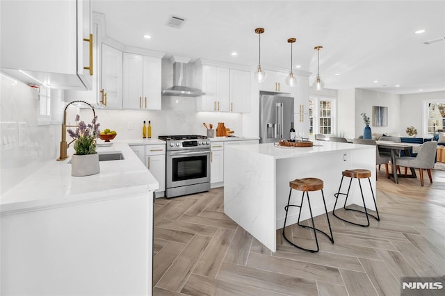 kitchen featuring a center island, wall chimney range hood, white cabinets, decorative light fixtures, and stainless steel appliances