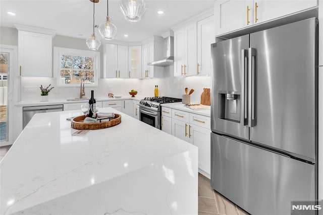 kitchen featuring wall chimney range hood, white cabinets, pendant lighting, stainless steel appliances, and light stone counters