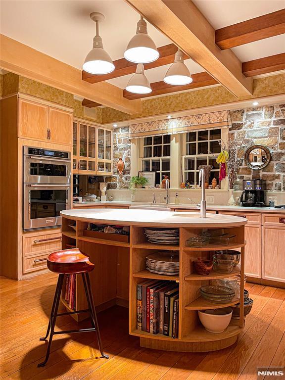 kitchen featuring double oven, hanging light fixtures, light hardwood / wood-style flooring, beam ceiling, and a breakfast bar area