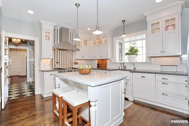 kitchen featuring a kitchen island, glass insert cabinets, a sink, wall chimney exhaust hood, and white cabinets